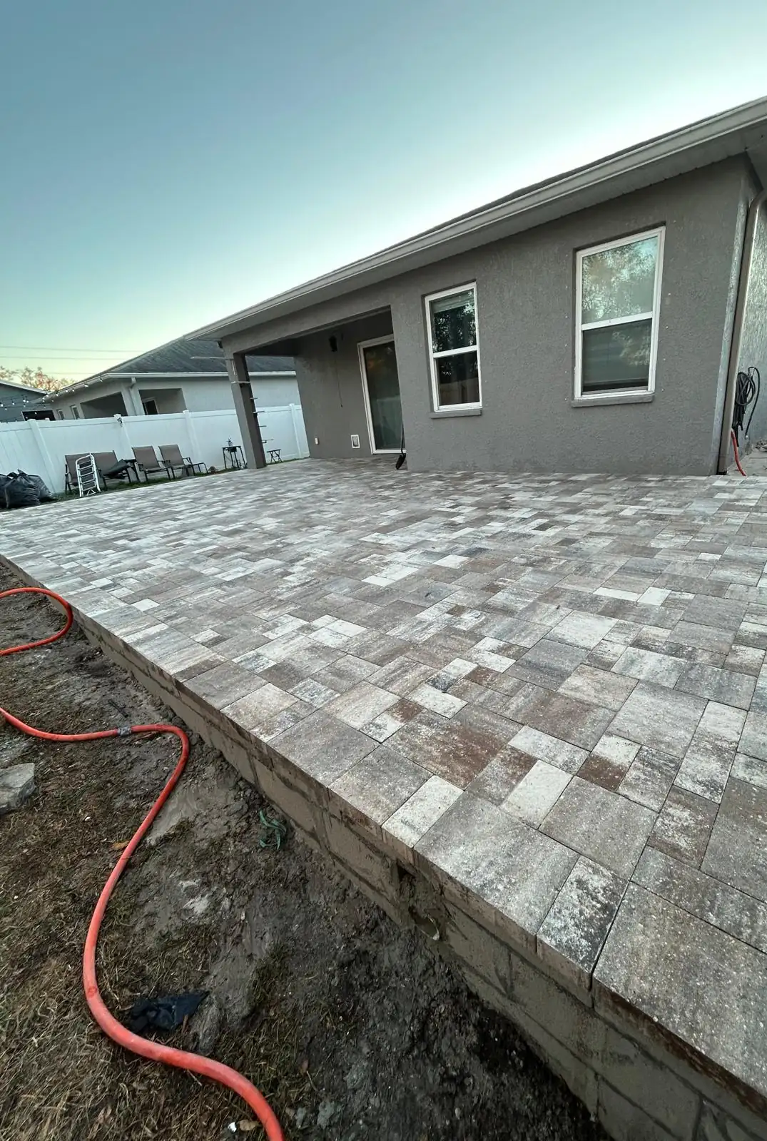 Completed paver patio along the back of the grey stucco house, raised on a concrete block edge.