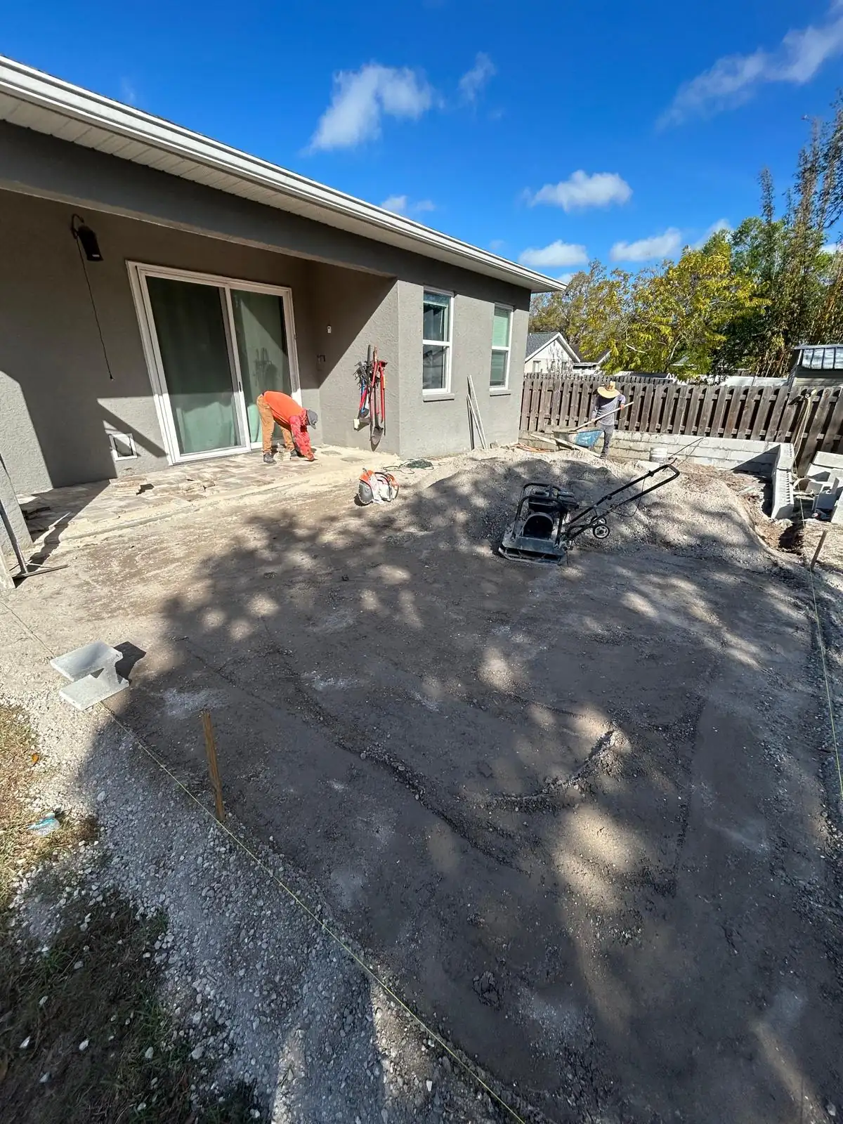 Crew running a plate compactor over the patio base behind the house before any pavers are set.
