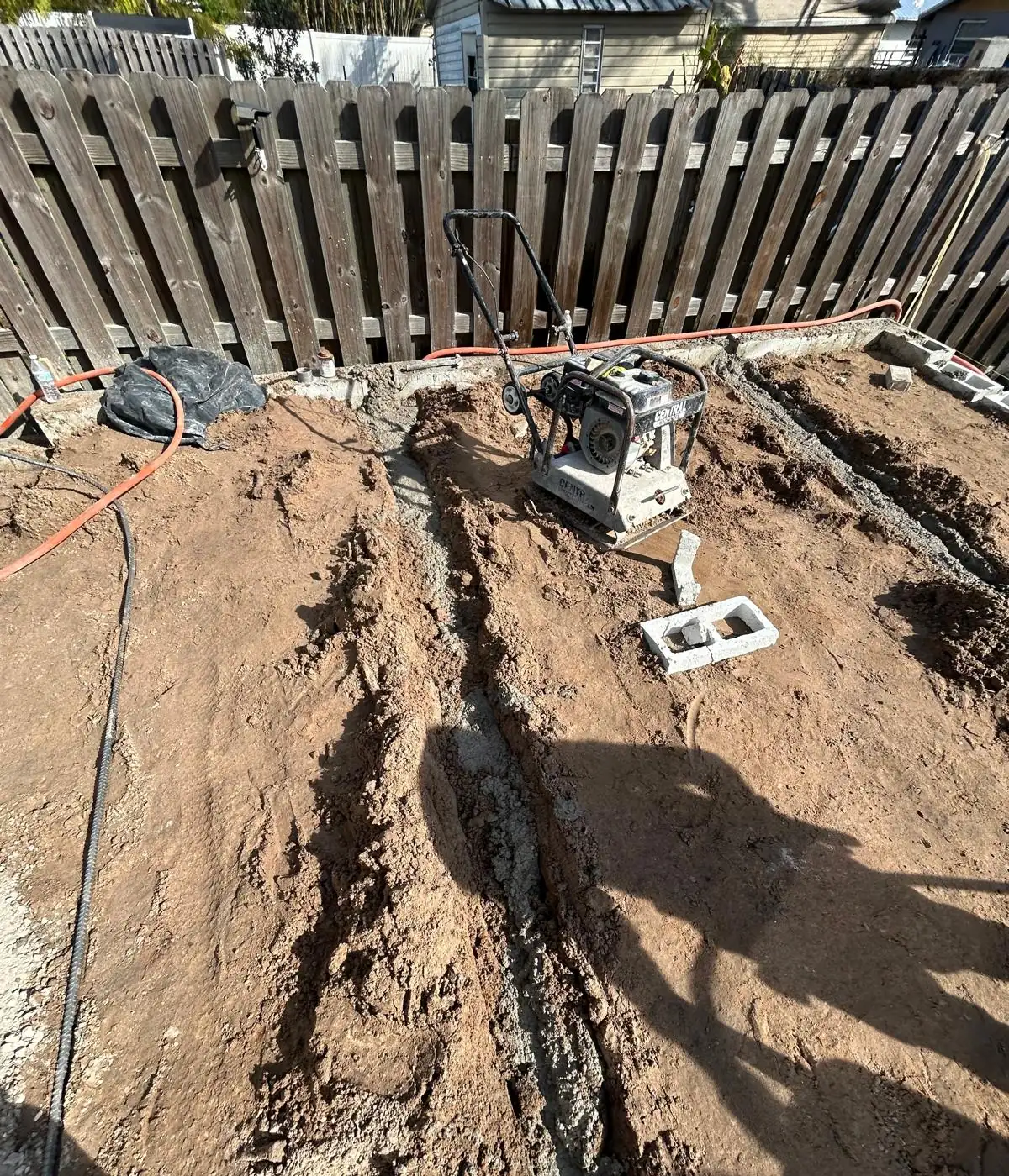 Plate compactor on the graded subgrade beside the concrete strip poured for the block edge along the fence.