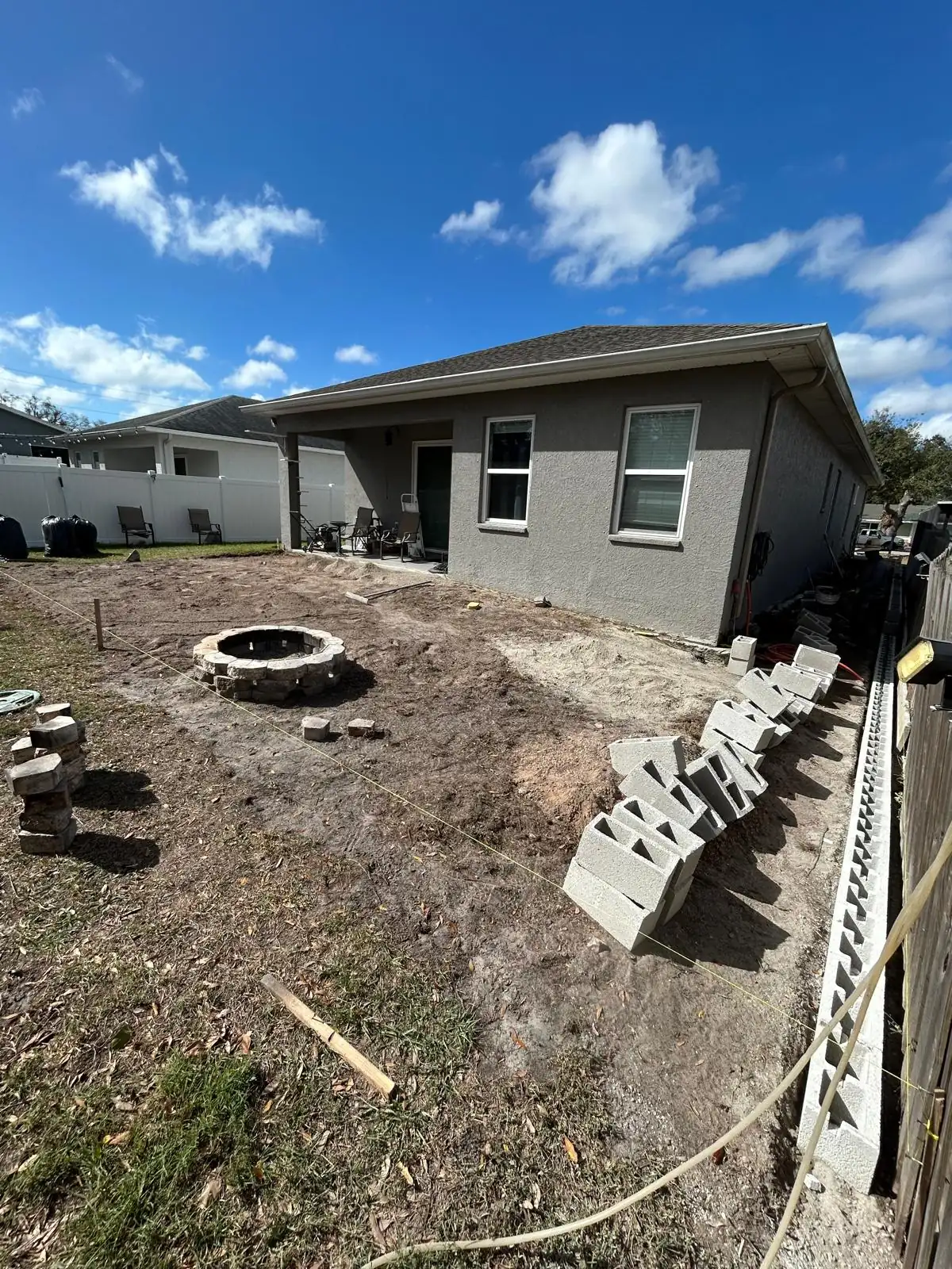 Bare dirt backyard of a grey stucco house before the patio, with a fire pit and staged concrete block.