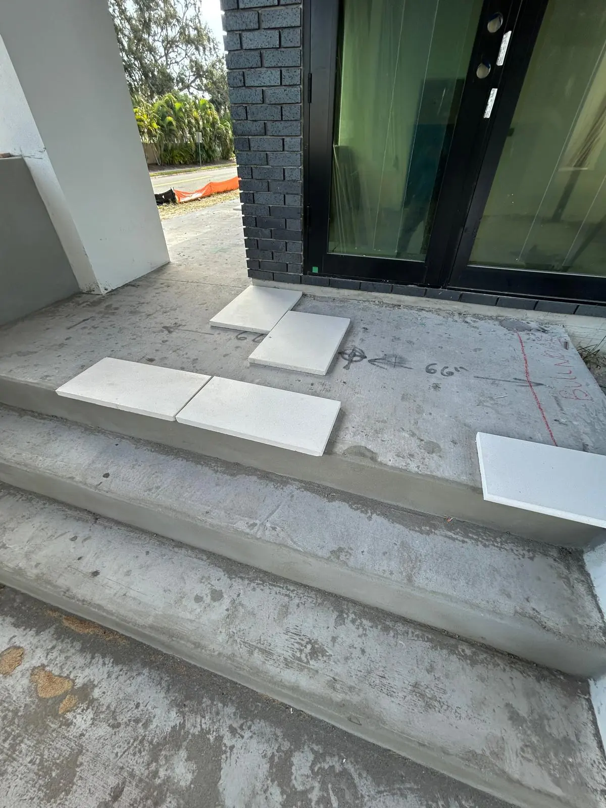 White stone slabs set out for layout on bare concrete entry steps below a dark brick and glass front door