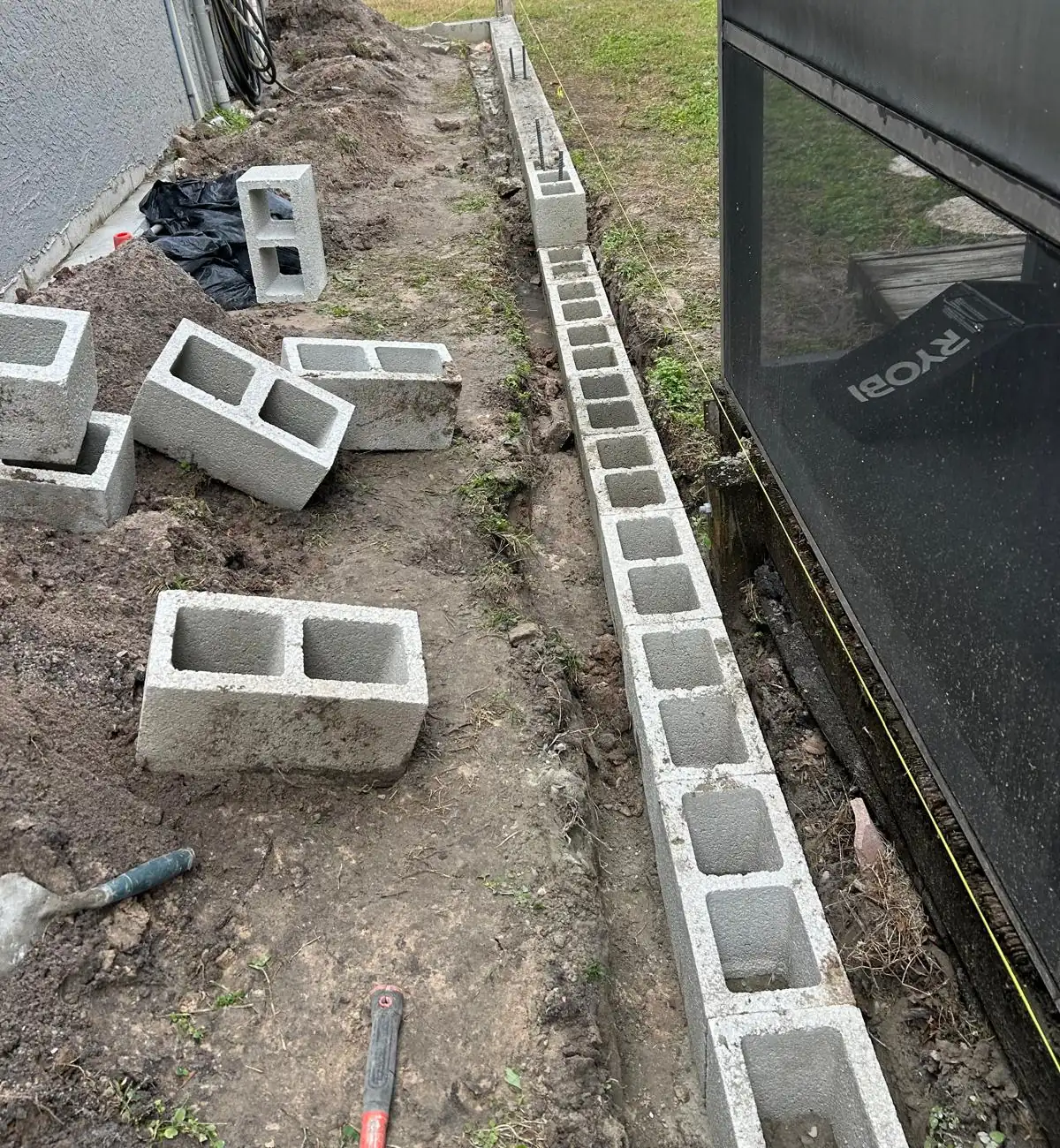 First course of concrete block set along the footer in the open trench beside a screen enclosure