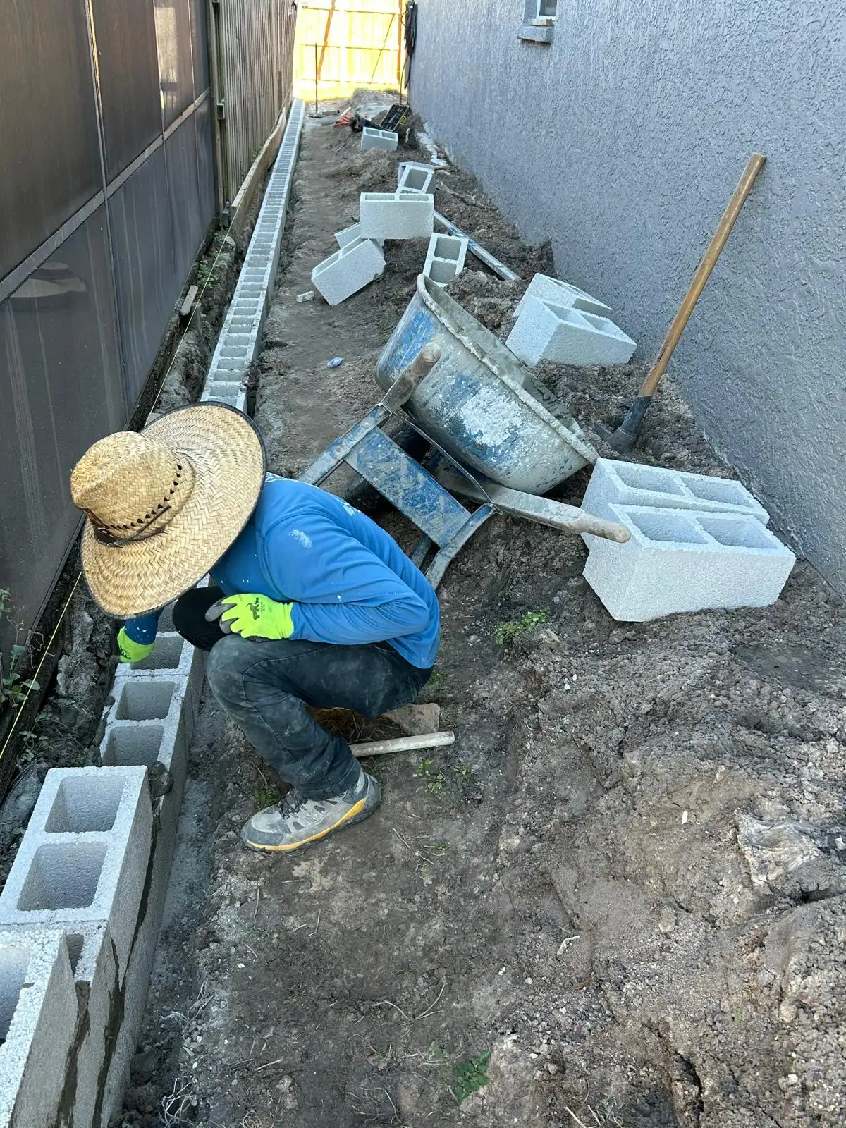 Worker setting block along the trench beside the screen enclosure, with a mortar tub and block staged nearby